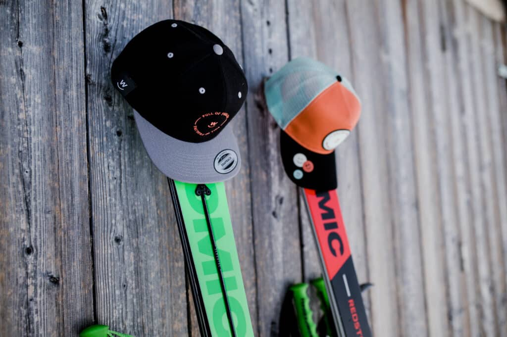2 Paar Ski lehnen an einer verwitterten Holzwand. Sie dienen jeweils für eine Baseballmütze als Kleiderständer